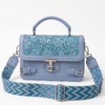 Dusty blue embossed floral faux leather top handle satchel bag with silver T-bar lock, buckle accents, and wide geometric woven shoulder strap.