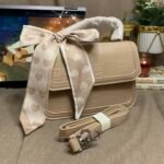 A beige embossed structured handbag sits on a rustic brown fabric surface next to a coiled shoulder strap wrapped in paper. In the blurred background are a laptop showing a cartoon, a potted plant with pink flowers, and a lit candle.