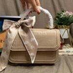 A woman's hand is shown holding a beige embossed handbag by its rigid top handle, which is currently encased in white protective paper wrapping. The decorative twilly scarf with hearts hangs below the handle. Blurry lifestyle background with a laptop and plant.