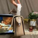 A structured beige handbag side profile view, held up to show the wide silver-tone metal zipper running down the side gusset. Blurry cozy lifestyle background. Scarf detail visible on top handle.