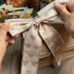Close-up of two hands expertly tying a beige twilly scarf, adorned with small heart patterns, into a perfect bow onto the rigid top handle of a beige structured bag. Laptop and plants are blurred in the background.
