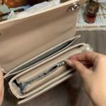 Inside view of an opened beige handbag. A hand pulls a central silver internal zipper. Multiple interior compartments and a fully lined interior are visible. Blurry lifestyle items in background.