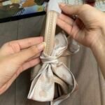 Hands are shown holding a beige structures satchel bag, pointing to the protective white paper wrapping that still encases the top handle, located just above the twilly scarf bow. Laptop blurred in background.