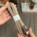 Hand holding a rolled-up, beige structured adjustable crossbody shoulder strap. It is wrapped with a protective white paper band and features durable silver-tone metal clasps at the ends. Cozie lifestyle background.